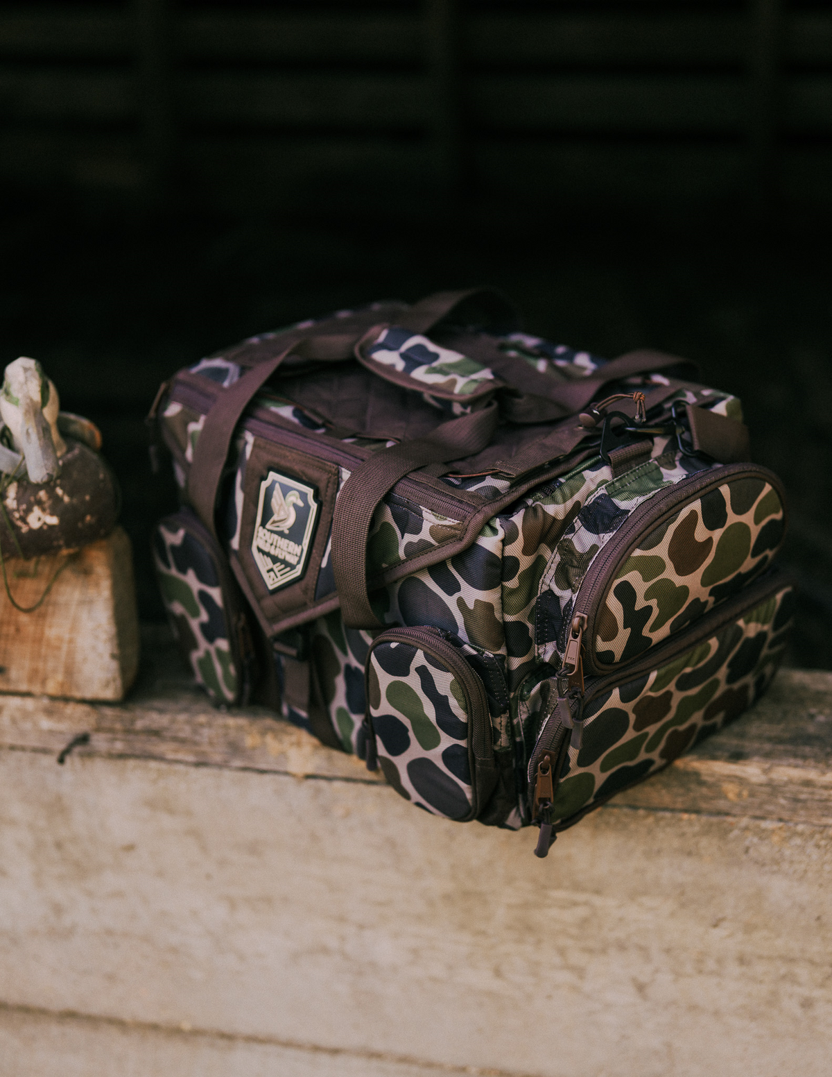 Large blind bag in vintage frogskin camo with the Deco Duck patch, next to a wooden duck decoy on a weathered ledge