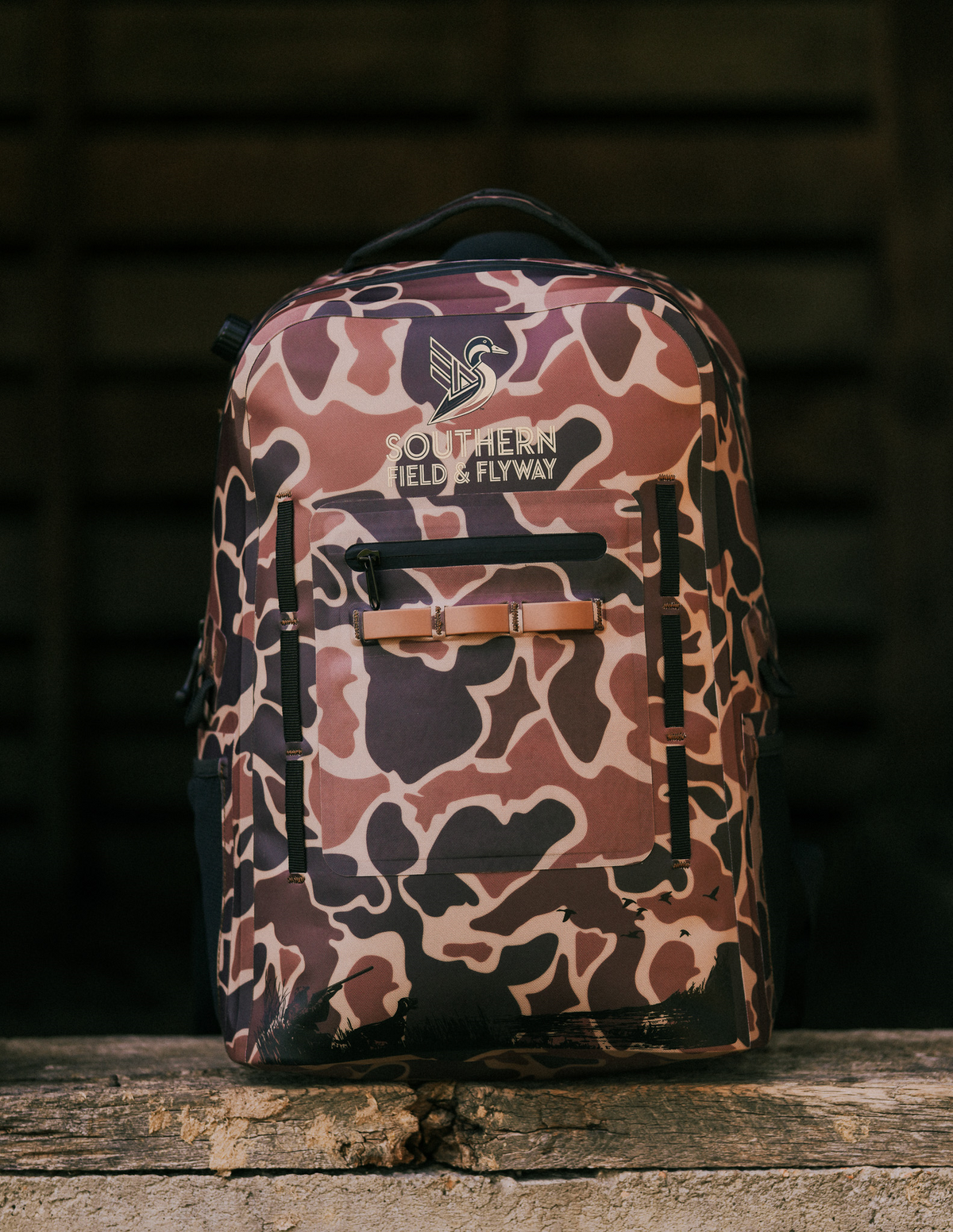Wetland waterproof hunting backpack in Marsh Camo with the Southern Field & Flyway logo, on a weathered wood ledge