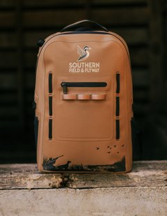 Wetland waterproof hunting backpack in Bayou Brown with the Southern Field & Flyway logo and a muddy base, on a weathered wood ledge